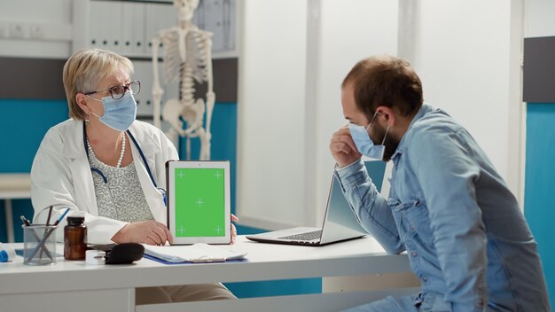 People with face mask using greenscreen at meeting in cabinet, on digital tablet. Man and specialist analyzing isolated chromakey background with mockup template and blank copyspace. Tripod shot.