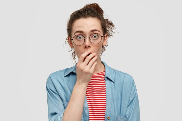 People and wonderment concept. Stunned freckled young female with frightened gaze, keeps hand on mouth, gossips with friend, gasps from fear and astonishment, wears round eyewear, casual shirt