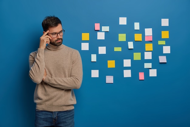 People, work, thoughts concept. contemplative bearded guy keeps finger on temple, looks pensively aside, puts colorful sticky notes on wall
