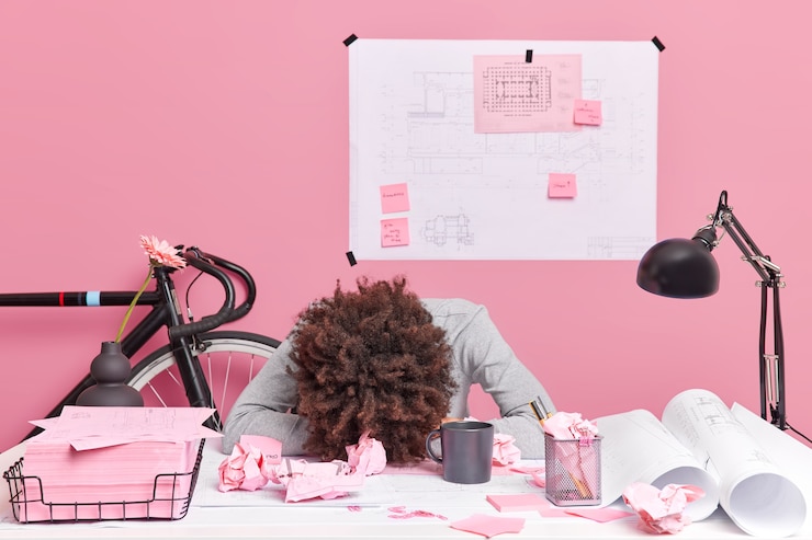 Free Photo people working exhaustion deadline concept. tired curly haired overworked woman leans at office desk works on future project surrounded with paper scraps sketches blueprints has lack of sleep