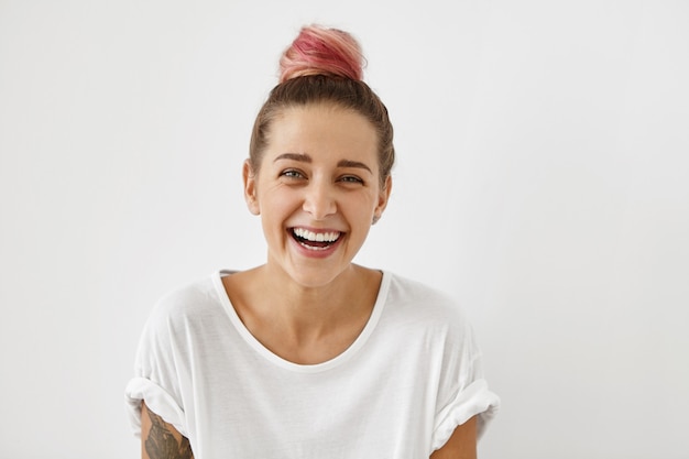 Free photo people, youth, joy, fun, happiness and relaxation concept. indoor portrait of trendy-looking hipster woman with hair knot dyed pink smiling cheerfuly, can't help but laughing while watching good comedy