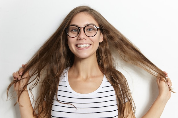 People, youth and lifestyle concept. funny cute adorable girl wearing trendy glasses and sailor shirt smiling excitedly, pulling her long hair, having fun indoors