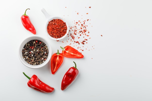 Peppercorns with chili powder in scoop and red peppers in a plate on white wall, top view.