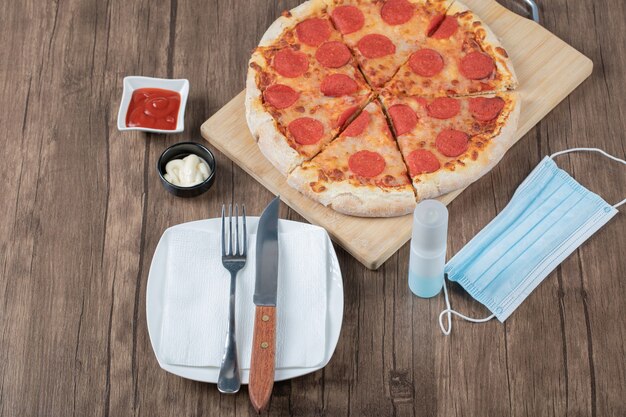 Pepperoni pizza on a wooden board with sauces, plate, hand sanitizer and mask around