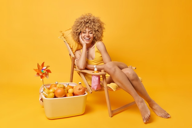 Free photo perfect summer holidays concept cheerful european woman with curly hair poses with glad expression on deck chair enjoys good rest uses portable cooler isolated over vivid yellow background