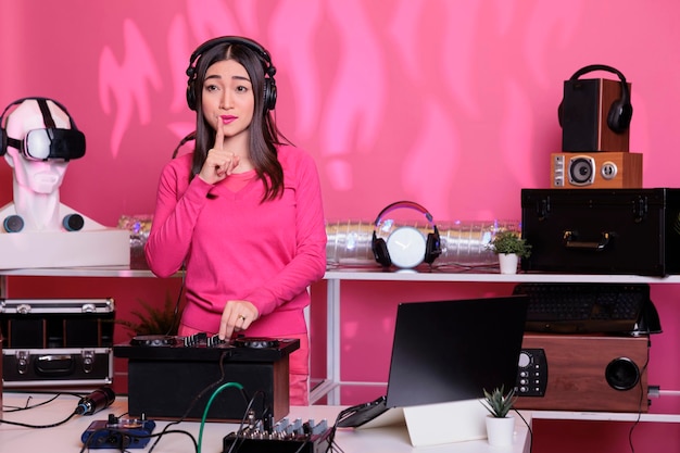 Free photo performer putting forefinger on lips gesturing secrecy sign while performing electronic music in studio over pink background. musical artist playing sounds to produce melody at mixer console