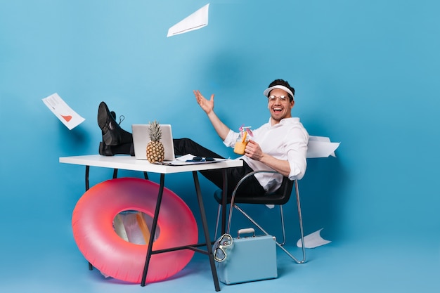 Perky businessman in suit and cap scatters graphics, enjoying tasty cocktail, sitting at table with laptop, rubber ring, suitcase on blue space.