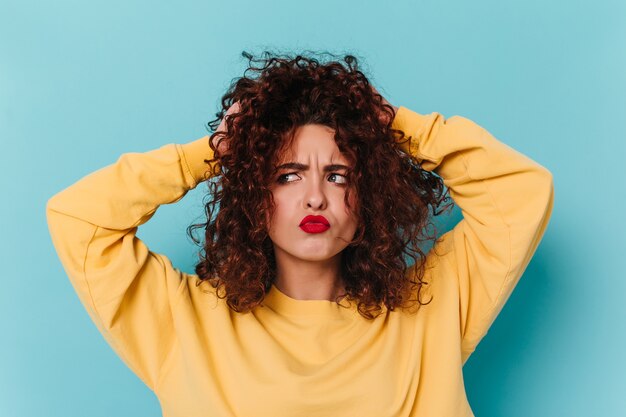 Perky girl musses up curly hair with funny facial expression. Shot of woman with red lips on blue space.