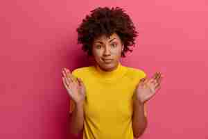 Бесплатное фото Озадаченная афроамериканка неуверенно поднимает ладони, не может помочь, стоит растерянно и нерешительно, говорит, что это не моя вина, носит желтый наряд, позирует над розовой стеной, пожимает плечами.