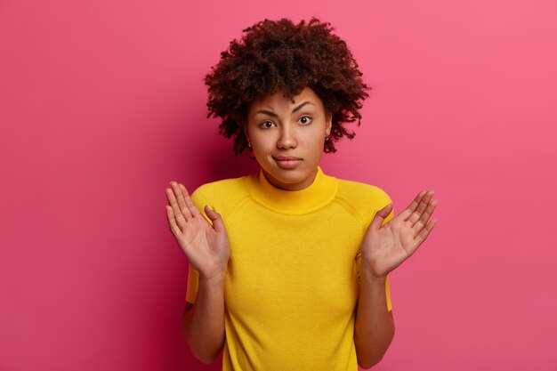 困惑したアフリカ系アメリカ人の女性は、ためらいがちに手のひらを上げ、助けができず、混乱して優柔不断に立ち、私のせいではないと言い、黄色い服を着て、ピンクの壁にポーズをとり、肩をすくめる