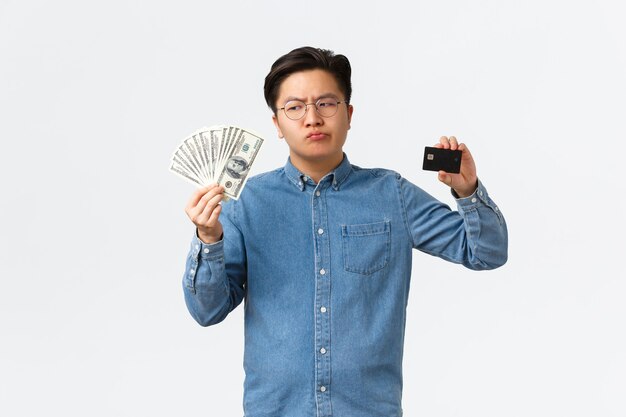 Perplexed asian businessman in glasses holding cash and money looking doubtful at money thinking use...