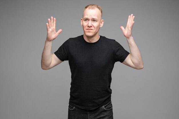 Озадаченный и грустный мужчина в черных рубашках поднимает руки вверх, позируя на сером фоне