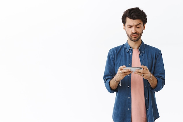 Perplexed thoughtful young handsome man thinking how pass difficult level smirk and frowning serious as looking smartphone screen hold mobile horizontally standing white background puzzled