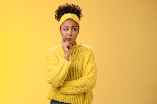 Perplexed unsure cute timid african-american female thinking how escape awkward situation smirking frowning concerned touch cheek thoughtful thinking nervously, worried yellow background.