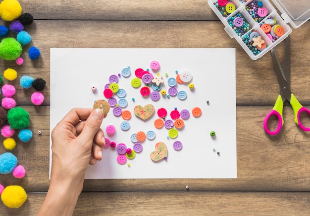 Free photo a person decorating white paper with colorful buttons over wooden table