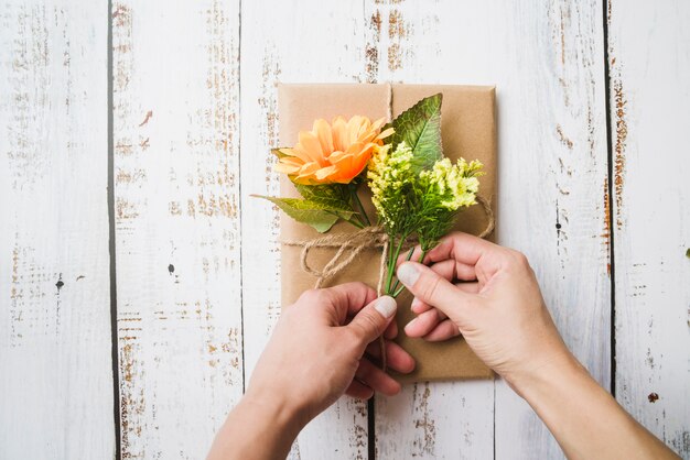 A person decorating the wrapped gift box with fake flowers on wooden background