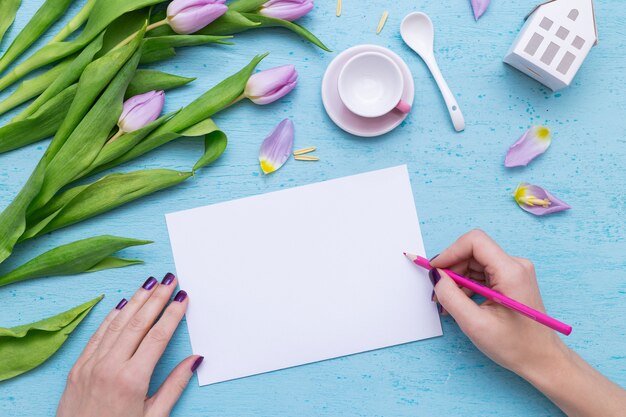 A person drawing on white paper with a pink pencil near purple tulips and a coffee cup