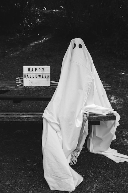 Free photo person in ghost costume sitting on bench near happy halloween inscription