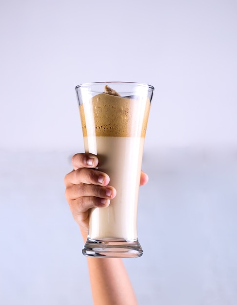 Person holding caramel smoothie in a glass behind a white wall