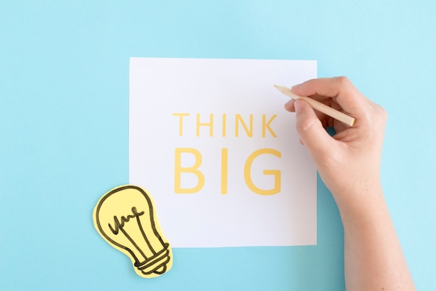 A person holding colored pencil written text think big on white paper over blue background