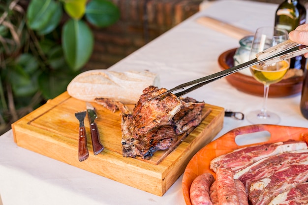 A person holding fresh grilled beef steak with tong on wooden board