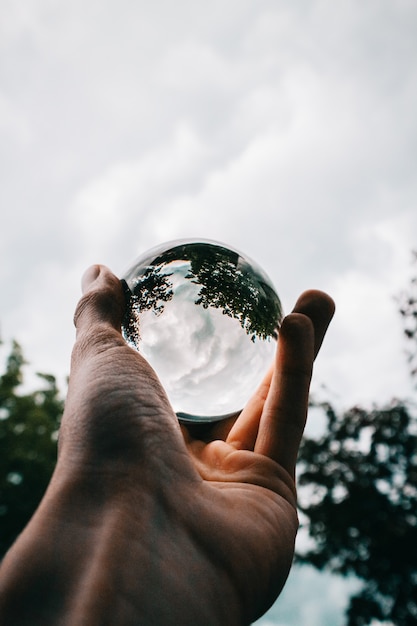 Free photo a person holding a glass ball with the reflection of beautiful green trees and breathtaking clouds