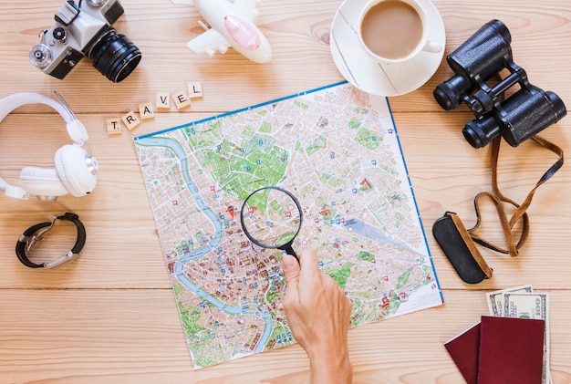 A person holding magnifying glass over map with tea cup and traveling equipment on wooden desk