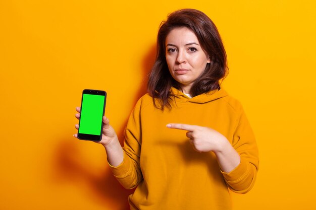 Человек, держащий мобильный телефон с вертикальным зеленым экраном в студии. Молодая женщина указывает на цифровое устройство с шаблоном макета и изолированным фоном на дисплее. Взрослый с мобильным телефоном