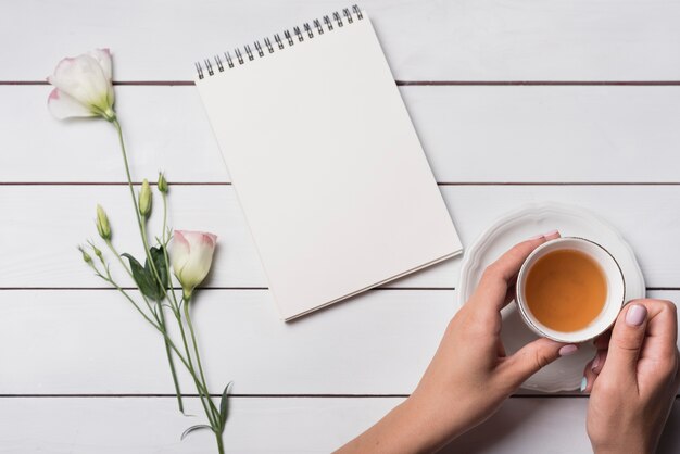 A person holding tea cup with spiral notepad and beautiful flowers on wooden desk