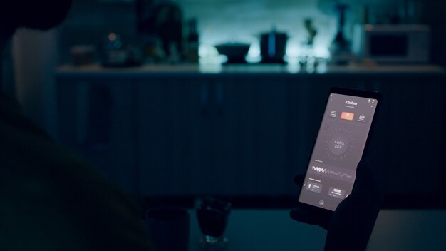 Человек смотрит на смартфон с приложением умного домашнего освещения, сидя на кухне дома с автоматизацией ...