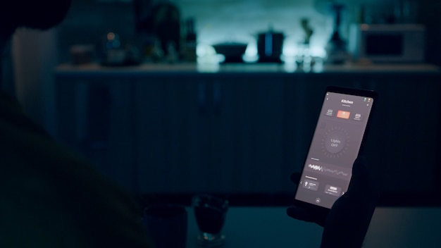 자동화된 집 부엌에 앉아 스마트 홈 조명 앱으로 스마트폰을 보고 있는 사람 ...