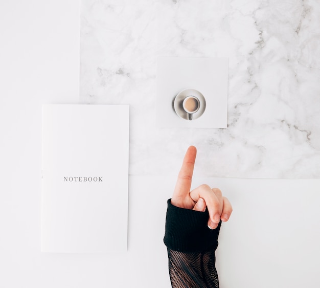 Free photo a person pointing finger to coffee cup with notebook on white desk