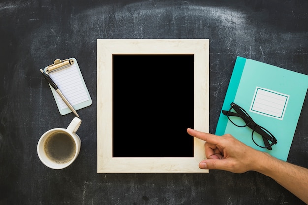 a person pointing finger on picture frame with stationeries and coffee cup on blackboard