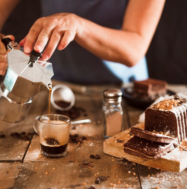 A person pouring espresso coffee in glass with cake slices on chopping board