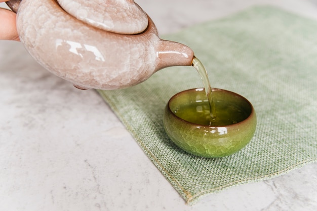 A person pouring herbal tea from the teapot in teacup on placemat