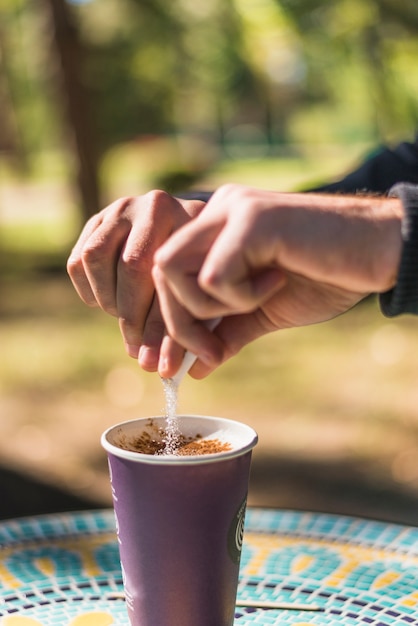 Рука человека, добавив сахар в чашку кофе на вынос на открытом воздухе