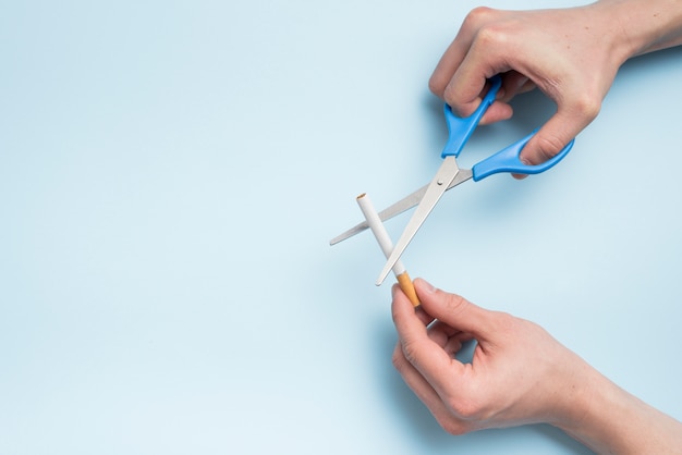 Person's hand cutting cigarette with scissor above blue background
