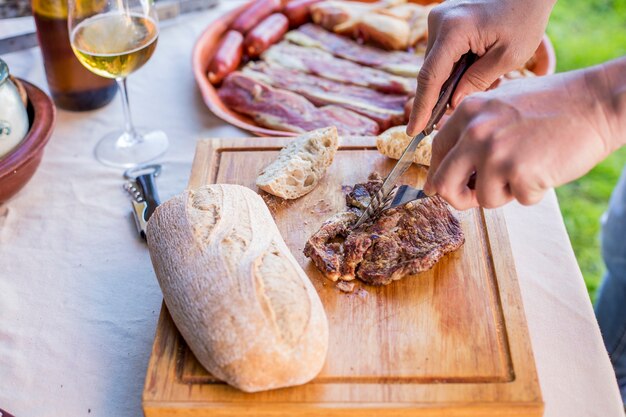 A person's hand cutting grilled beef steak on chopping board with knife and fork