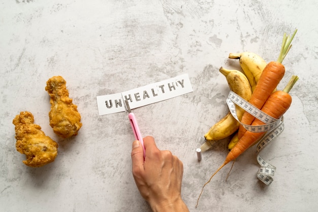 Person's hand cutting unhealthy word near fried chicken with banana and carrots rolled in measuring tape