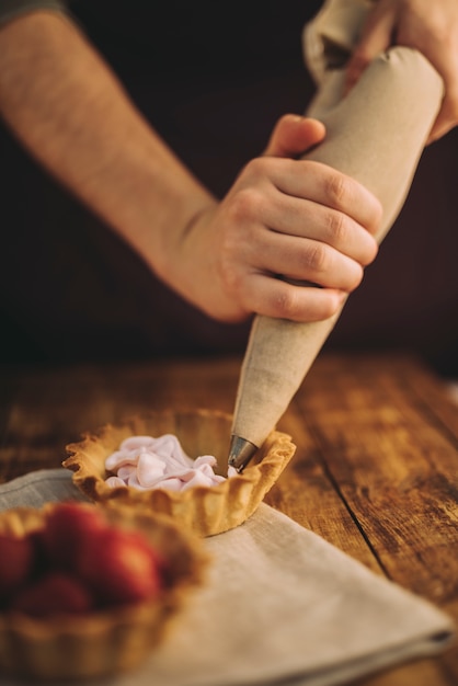 A person's hand filling the tart with pink whipped cream from icing bag on wooden table