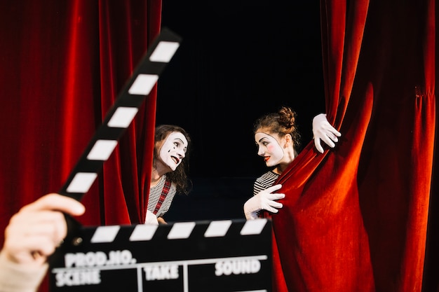 A person's hand holding clapperboard in front of two mime artist performing behind red curtain