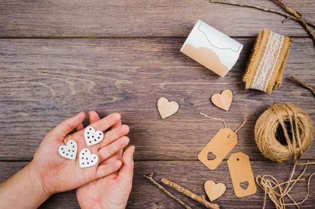 A person's hand holding heart shape with jute spool; tags; sticks and lace on wooden desk