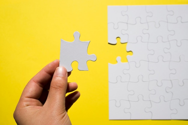 A person's hand holding missing white jigsaw puzzle piece over yellow backdrop