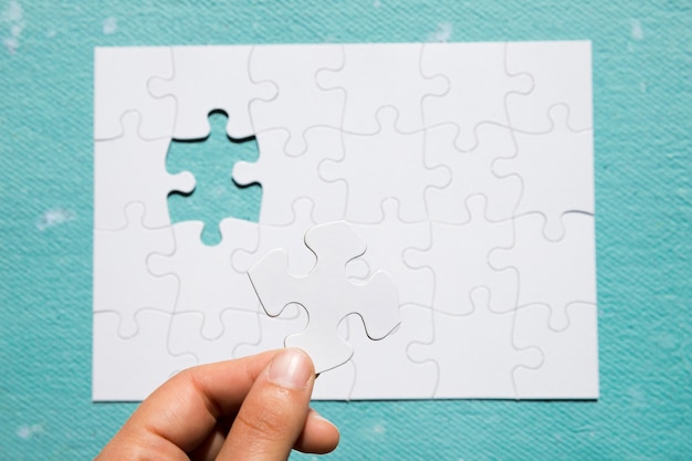 Free photo a person's hand holding white puzzle piece on puzzle grid over blue textured background