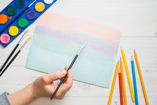 Person's hand painting on paper with paint brush and watercolor over desk