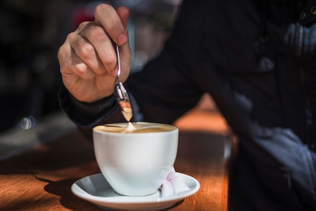 A person's hand stirring latte coffee with spoon in white coffee cup on wooden table