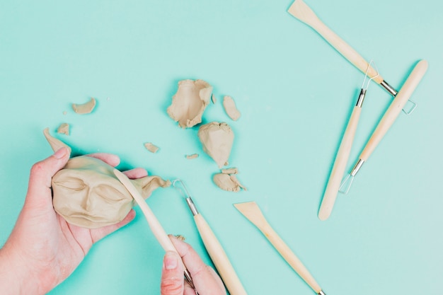 A person's hand using sculpting tools for making clay face on mint green backdrop