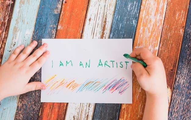 Person's hand writing on white paper with green crayon over weathered wooden desk