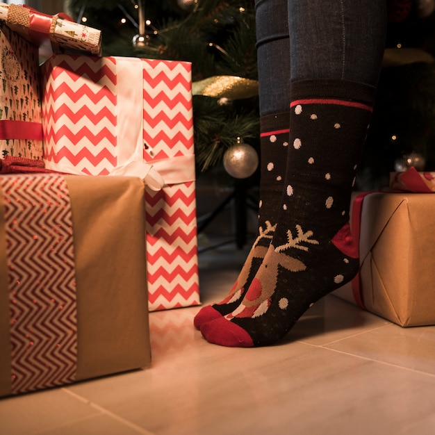 Person's legs in Christmas socks between present boxes and decorated fir tree