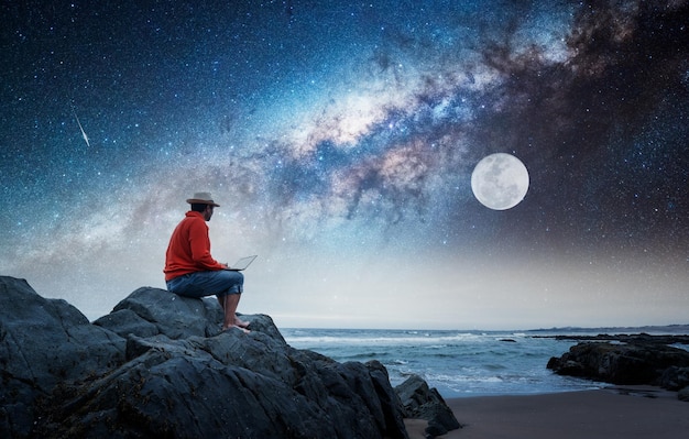 月​と​天の川​を​見つめている​ビーチ​で​ラップ​トップ​を​持って​岩​の​上に​座っている​人